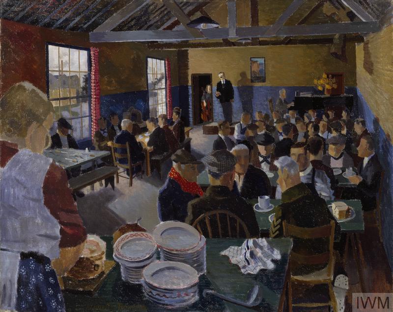 The interior of a large room, filled with people seated around tables.  In the foreground there is a table stacked with plates, behind which stands a woman wearing an apron. 